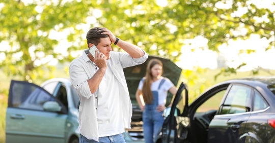 Man on phone with a car accident scene behind him.