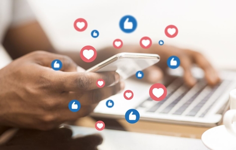 Using laptop and holding phone. Like and love emojis flutter around the phone